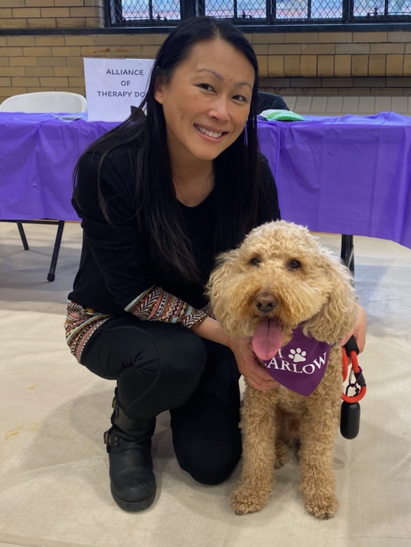 An Asian woman with long hair dressed in black crouched down next to a medium sized apricot colored mini labradoodle